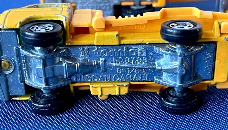 May include: Close-up of a yellow and blue toy truck. The underside of the truck is blue with black wheels. The text "Tomica NO.87,88" and "NISSAN CABALL" are visible.