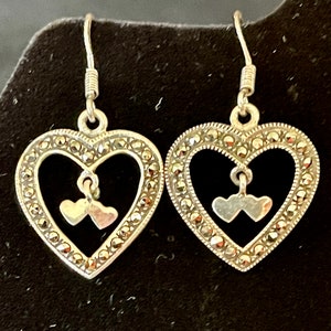 May include: A pair of silver dangle earrings with a heart design. The hearts are made of marcasite and have a smaller heart charm hanging from the centre.