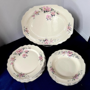 May include: A collection of off-white ceramic serving dishes with a pink floral pattern and a black trim. The set includes an oval platter, several oval plates, and a stack of bowls. The dishes are arranged on a dark blue surface.
