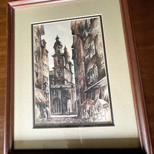 Könnte beinhalten: Eine Schwarzweiß-Tuschzeichnung einer engen Straße in einer europäischen Stadt. Die Straße ist von hohen Gebäuden gesäumt, und im Hintergrund befindet sich ein Kirchturm. Die Zeichnung ist in einem braunen Holzrahmen gerahmt.