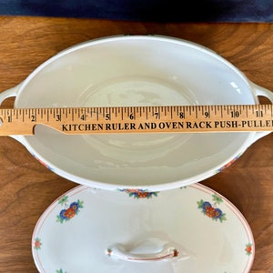 May include: White oval ceramic serving dish with a floral pattern in red, blue, and green. The dish has two handles and measures approximately 10 inches in length. The dish is accompanied by a matching lid with a floral pattern.