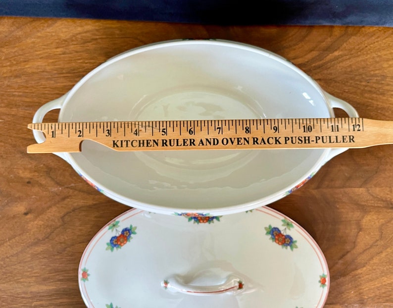 May include: White oval ceramic serving dish with a floral pattern in red, blue, and green. The dish has two handles and measures approximately 10 inches in length. The dish is accompanied by a matching lid with a floral pattern.