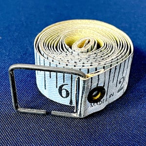 Könnte beinhalten: Ein aufgerolltes, hellblaues Maßband mit schwarzen Markierungen und der Zahl 6 in Schwarz. Das Band hat ein Metallendstück und die Worte "MADE IN" sind sichtbar. Das Band misst bis zu 15 cm.