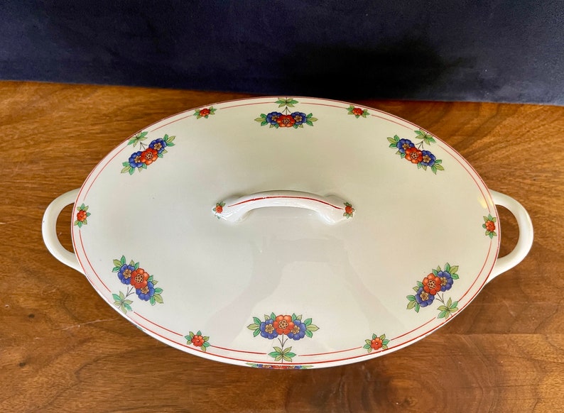 May include: White oval serving dish with a floral design in red, blue, and green. The dish has two handles and a lid with a handle.