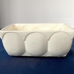 Puede incluir: Una jardinera rectangular de cerámica blanquecina con acabado moteado. Los lados presentan tres diseños grandes, redondeados y en relieve. El interior es de un tono más claro de blanquecino.