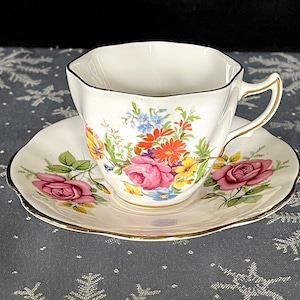 Op de afbeelding: Een wit theekopje en schotel met een bloemenpatroon. Het patroon heeft roze, gele, rode en blauwe bloemen. Het kopje en de schotel hebben een gouden rand.
