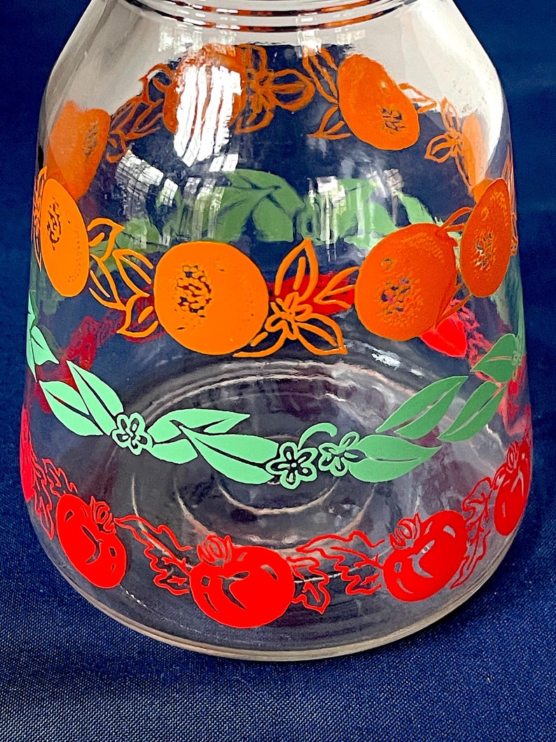 May include: Clear glass pitcher with a wide base and a narrow neck. The pitcher is decorated with orange, green, and red fruit and floral designs. The fruit appears to be oranges and tomatoes. The pitcher is likely vintage.
