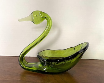 May include: A green glass swan shaped dish. The swan's neck curves up and the dish is shaped like a swan's body.