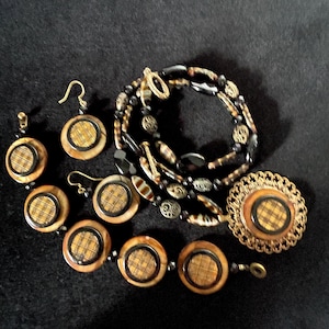 Puede incluir: Una colección de joyas que incluye una pulsera, un collar, aretes y un broche. Las piezas presentan un diseño circular con un patrón de cuadros en el centro, rodeado por un borde oscuro y un marco marrón claro. El collar y la pulsera tienen cuentas negras.