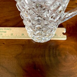Op de afbeelding: Een glazen kan met een diamantpatroon. De kan heeft een handvat en staat op een houten oppervlak.