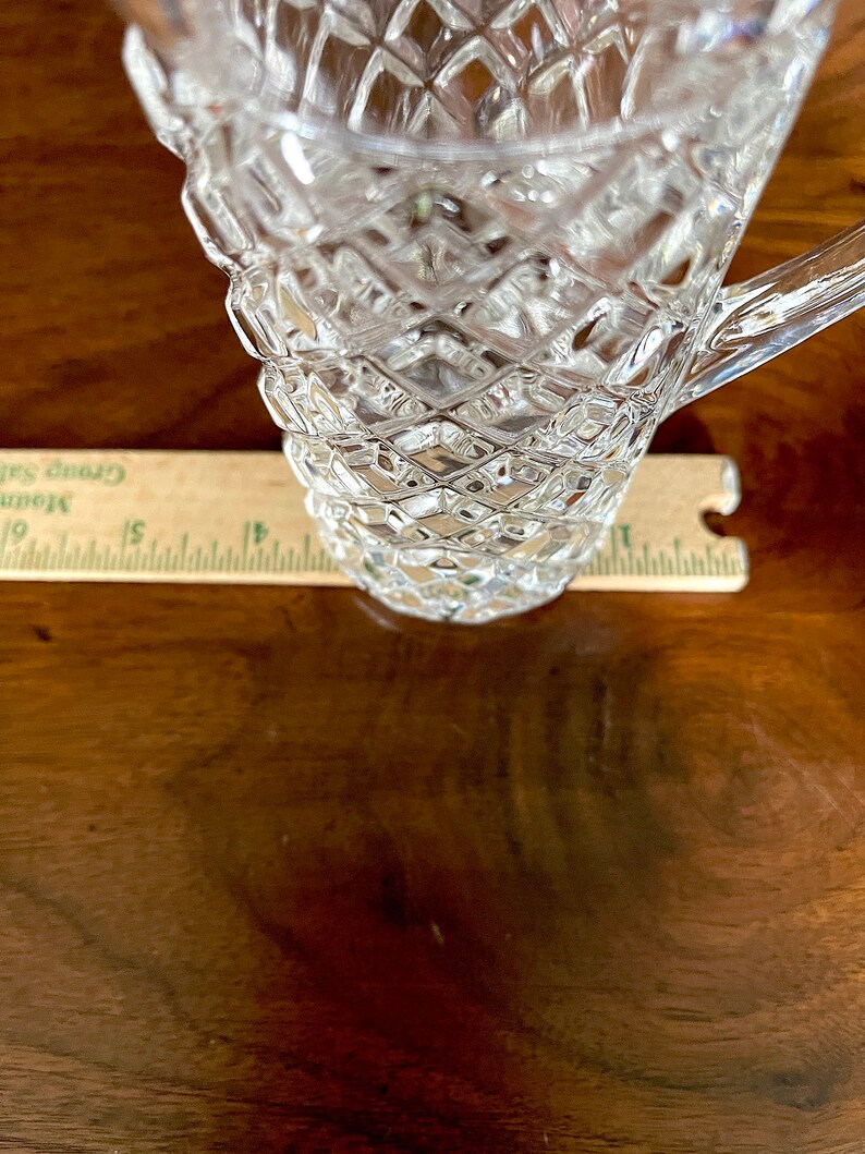Op de afbeelding: Een glazen kan met een diamantpatroon. De kan heeft een handvat en staat op een houten oppervlak.