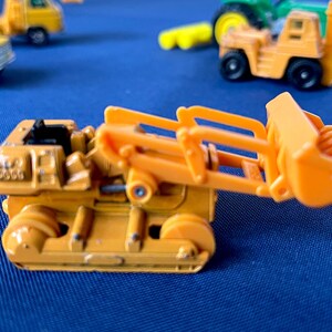 May include: An orange toy bulldozer with a raised bucket, set against a blue background. Other toy construction vehicles, including a yellow truck and a forklift, are visible in the background. The bulldozer has black wheels and detailed features.