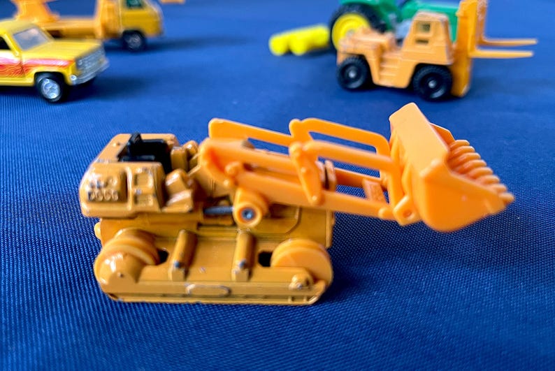 May include: An orange toy bulldozer with a raised bucket, set against a blue background. Other toy construction vehicles, including a yellow truck and a forklift, are visible in the background. The bulldozer has black wheels and detailed features.