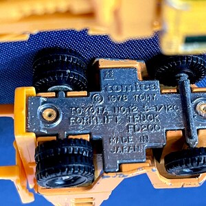May include: A close-up of a yellow toy forklift truck with black wheels. The underside of the toy is visible, showing the text "comica &copy; 1978 TOMY TOYOTA NO.12 6-1/120 FORKLIFT TRUCK FD200 MADE IN JAPAN."