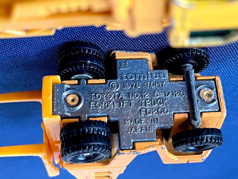 May include: A close-up of a yellow toy forklift truck with black wheels. The underside of the toy is visible, showing the text "comica &copy; 1978 TOMY TOYOTA NO.12 6-1/120 FORKLIFT TRUCK FD200 MADE IN JAPAN."