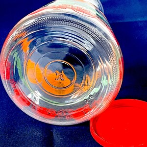 May include: Clear glass jar with a red lid, featuring colorful fruit designs around the rim. The jar's base has an embossed design. The lid is bright red, and the jar is set against a dark blue background.