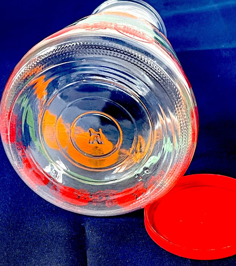 May include: Clear glass jar with a red lid, featuring colorful fruit designs around the rim. The jar's base has an embossed design. The lid is bright red, and the jar is set against a dark blue background.