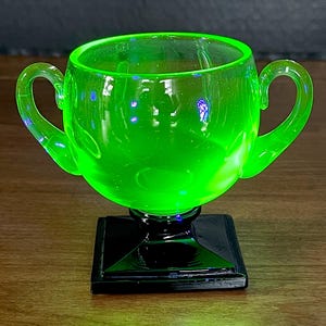 May include: A vibrant neon green glass cup featuring two curved handles on the sides. The cup is supported by a solid black square base and a short pedestal, creating a distinct contrast between the bright translucent bowl and the dark foundation.