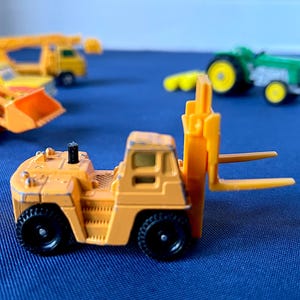 May include: A small, orange toy forklift with black wheels and a yellow fork. Other toy construction vehicles are in the background, including a yellow truck and a green tractor. The toys are on a blue surface.