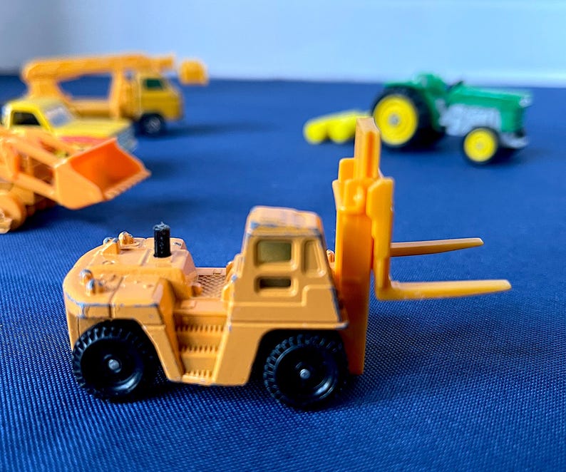 May include: A small, orange toy forklift with black wheels and a yellow fork. Other toy construction vehicles are in the background, including a yellow truck and a green tractor. The toys are on a blue surface.