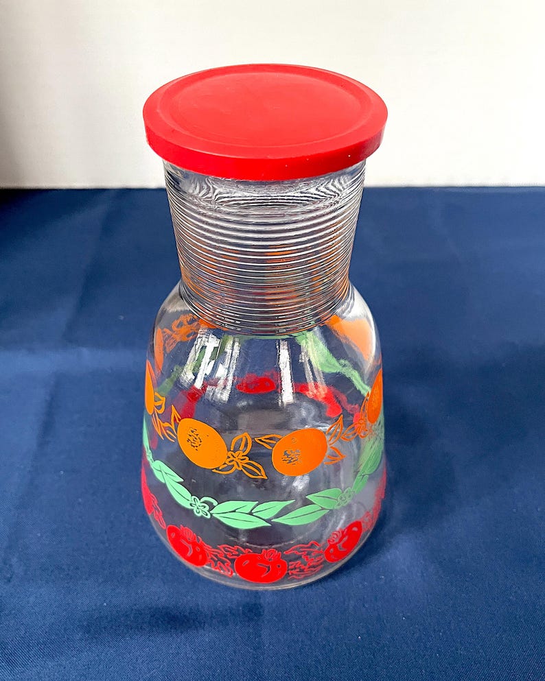 May include: A clear glass pitcher with a red lid. The pitcher is decorated with orange and red fruit designs and green leaves. The pitcher has a ribbed neck and a rounded body. A vintage kitchenware item.