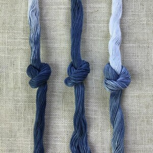 Può includere: Tre matassine di filo da ricamo con una sfumatura di colore blu. Ogni filo è annodato al centro, passando da un azzurro chiaro o bianco nella parte superiore a un blu navy profondo e saturo verso le estremità inferiori dei fili.