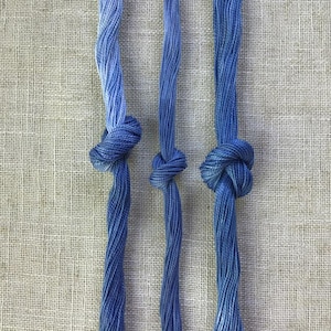 Può includere: Tre fili di cotone da ricamo blu disposti verticalmente, ciascuno annodato con un semplice nodo. I fili presentano un motivo cromatico variegato, che sfuma da tonalità azzurro cielo chiaro a blu navy intenso lungo la loro lunghezza.