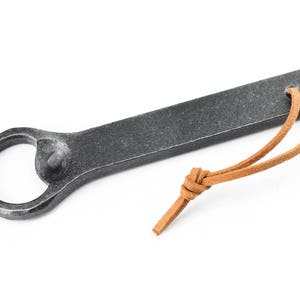 A grey blacksmith made iron bottle opener with a light brown leather tassel on a white background.