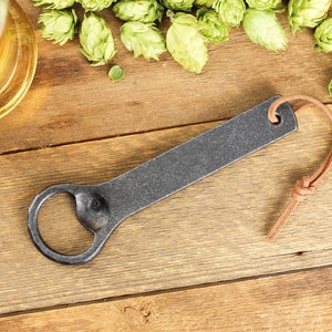 A handmade iron bottle opener with a light brown leather tassel rests on a rustic barn board surface surrounded by fresh green hops and a tall glass of beer.
