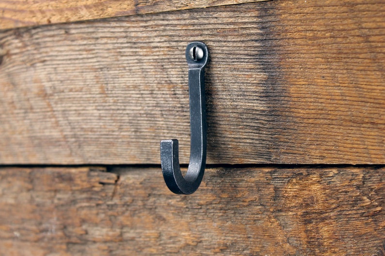 May include: A black metal hook is mounted on a wooden surface. The hook is made of forged metal and has a simple, rustic design.