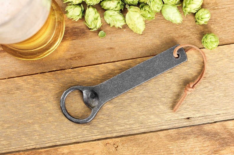 A handmade iron bottle opener with a light brown leather tassel rests on a rustic barn board surface surrounded by fresh green hops and a tall glass of beer.