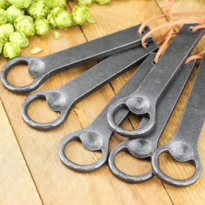 A group of hand forged iron bottle openers laid on a rustic barn board tabletop. Fresh green hops in the background.