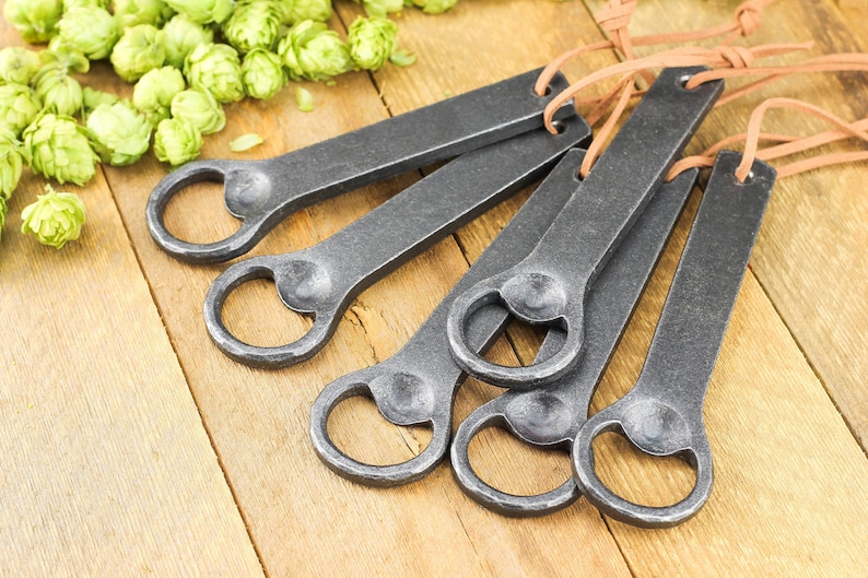 A group of hand forged iron bottle openers laid on a rustic barn board tabletop. Fresh green hops in the background.