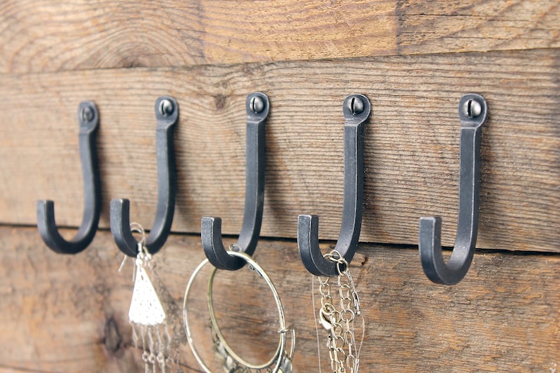 May include: Five black metal hooks mounted on a wooden wall. The hooks are in a row and have a simple J-shape. Jewelry is hanging from some of the hooks.