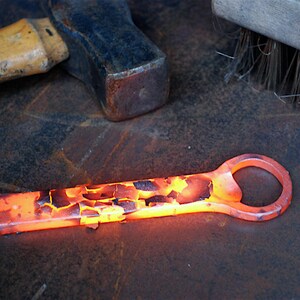 A glowing hot bottle opener rests on a rustic metal workbench after being forged. Oxidized metal scale flakes from the surface of the handle. A blacksmiths hammer and a wire brush are in the background.