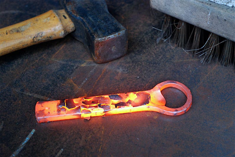 A glowing hot bottle opener rests on a rustic metal workbench after being forged. Oxidized metal scale flakes from the surface of the handle. A blacksmiths hammer and a wire brush are in the background.