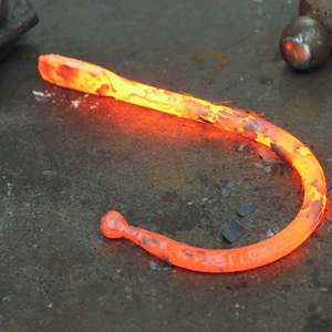 Blacksmith made iron curtain hook, glowing hot on a rustic metal workbench.