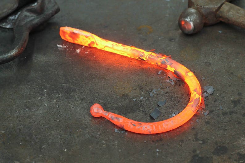 Blacksmith made iron curtain hook, glowing hot on a rustic metal workbench.