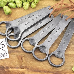 Multiple forged iron bottle openers are arranged on a rustic wood surface, surrounded by bright green hops.