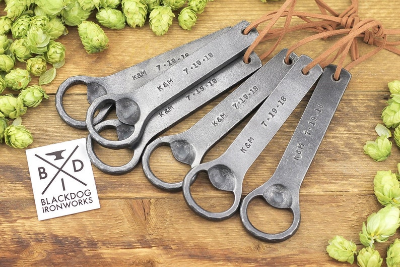 Multiple forged iron bottle openers are arranged on a rustic wood surface, surrounded by bright green hops.