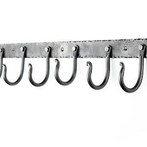 Könnte beinhalten: Ein schmiedeeiserner, an der Wand montierter Kleiderhaken mit sechs gebogenen Haken. Der Haken besteht aus dunkelgrauem Metall und hat eine rechteckige Stange mit Nieten. Die Haken sind gleichmäßig verteilt und haben ein dekoratives, geschwungenes Design.