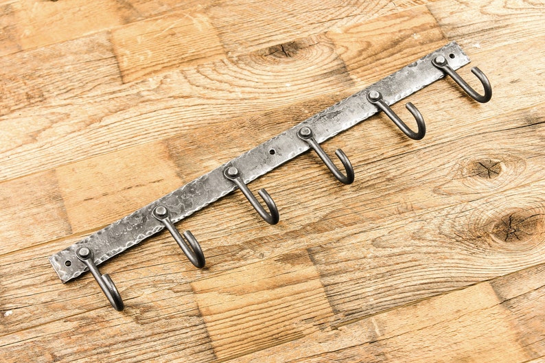May include: A metal wall-mounted hook rack with six hooks. The rack is made of hammered metal and has a rustic finish. The hooks are curved and are ideal for hanging keys, coats, or other items.