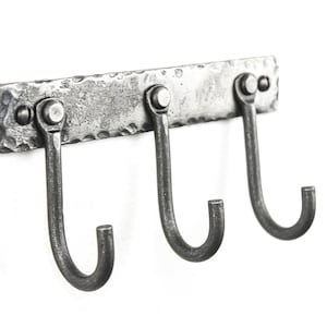 May include: A silver metal wall mounted hook rack with three hooks. The rack has a hammered texture and is mounted with three screws.