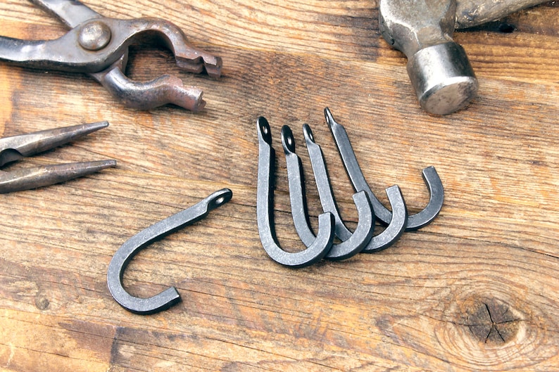 May include: Five black metal hooks on a wooden surface. The hooks are handmade and have a rustic look. There are also two metal tools on the surface, including a hammer and pliers.