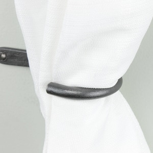 Puede incluir: Un amarre de cortina de metal negro con un gancho curvo. El amarre está montado en una pared y sujeta una cortina blanca.