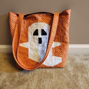 Op de afbeelding: Oranje en witte quilted tote bag met een spookontwerp. Het spook is wit met zwarte ogen en een mond.