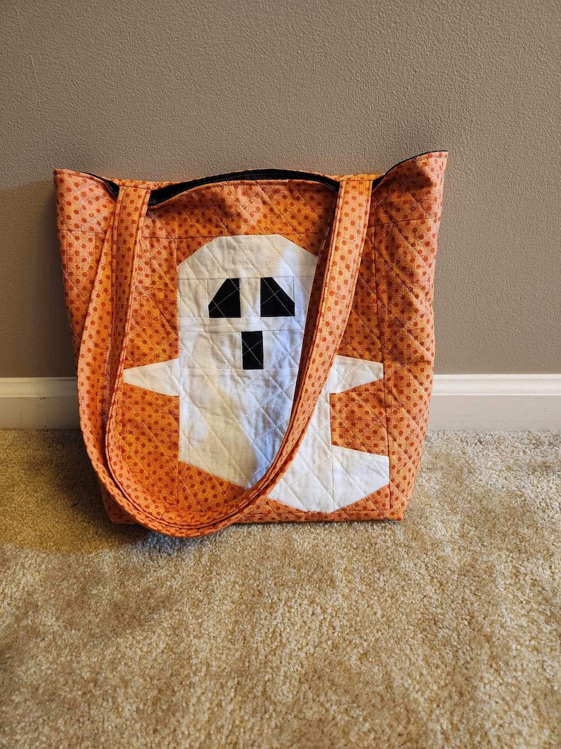 Op de afbeelding: Oranje en witte quilted tote bag met een spookontwerp. Het spook is wit met zwarte ogen en een mond.
