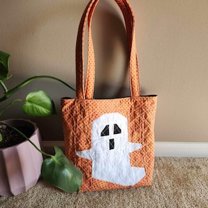 Op de afbeelding: Oranje en witte quilted tote bag met een spookontwerp. De tas heeft zwarte ogen en een mond.
