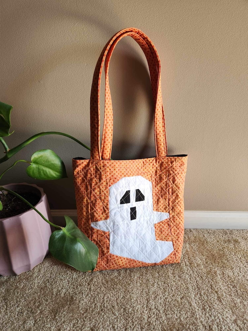 Op de afbeelding: Oranje en witte quilted tote bag met een spookontwerp. De tas heeft zwarte ogen en een mond.