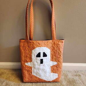 Op de afbeelding: Oranje en witte Halloween-tas met een spookontwerp. De tas heeft een wit spook met zwarte ogen en een mond op een oranje achtergrond met witte stippen.